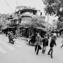 A black and white street photography of Hanoi streets in Vietnam, with people crossing the road at crosswalks and pedestrians on both sides of them, surrounded by old buildings, cars, motorbikes, bicycles, trees, greenery, bustling crowds, and children playing happily. It is a wide shot.