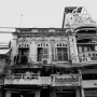 A black and white photo of the old architecture in Hanoi, Vietnam. The building has an ornate facade with arched windows and decorative elements like wooden shutters or balcony railings. It is surrounded by other buildings, some tall with modern glass facades, others more traditional with woodwork and tiles. There are lots of wires and cables hanging from above, adding to its urban atmosphere. In front there are street food stalls selling typical Vietnamese dishes such as pho and banh mi.