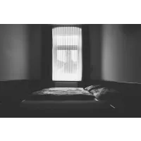 A simple, monochrome photograph of an empty bed in front of the window, symbolizing sleepless nights and loneliness. The dimly lit room has soft shadows that add to its melancholic atmosphere. Offering comfort but no real solace for those who have lost someone or something during their night.