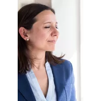 A portrait of an adult woman in her late thirties, wearing professional attire with a blue blazer and white shirt, with medium brown hair, eyes closed smiling slightly, standing by the window, with soft natural light, with a shallow depth of field. Capturing her confident expression and friendly demeanor in the style of a realistic portrait.